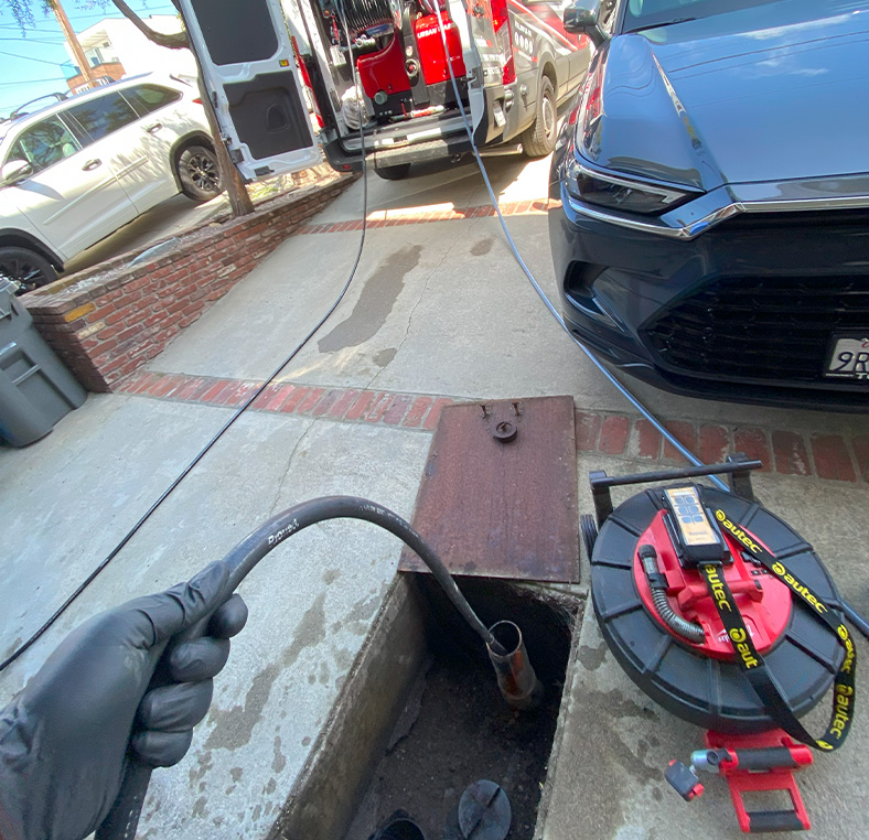 Plumber feeding a drain snake cable into an outdoor sewer cleanout on a residential driveway