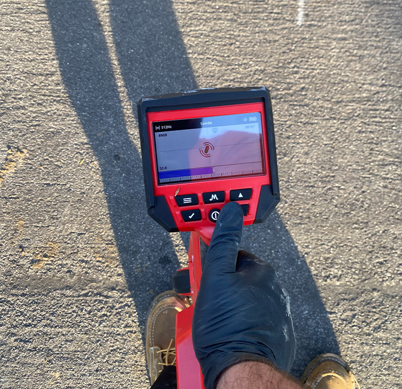 Gloved hand holding a red handheld sewer inspection camera monitor on pavement