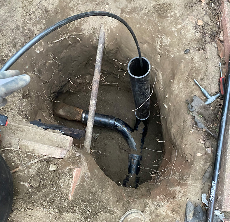 Excavated trench revealing a damaged lateral and a new vertical replacement pipe under concrete