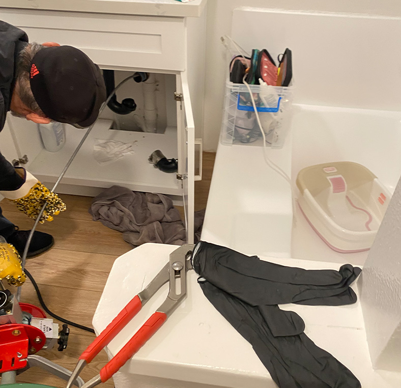 Indoor bathroom plumbing repair under a bathtub using a drain snake and wrenches